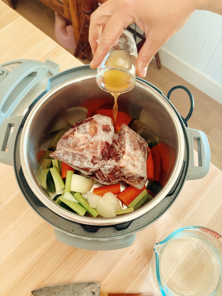 The instant pot contains the beef bones and roughly chopped vegetables. They are now adding the apple cider vinegar into the instant pot as well.