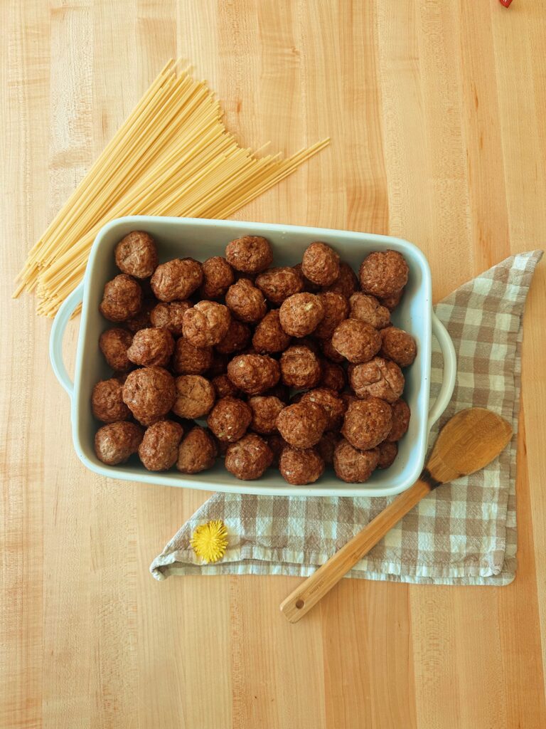 "Discover Your Next Dinner" This image is of  a large amount of meatballs in a glass casserole dish. There are dry pasta noodles, a wooden spoon, a dandelion, and a brown gingham napkin on the butcher block a well. This link will take us to the recipe index with recipe ideas for dinners. 