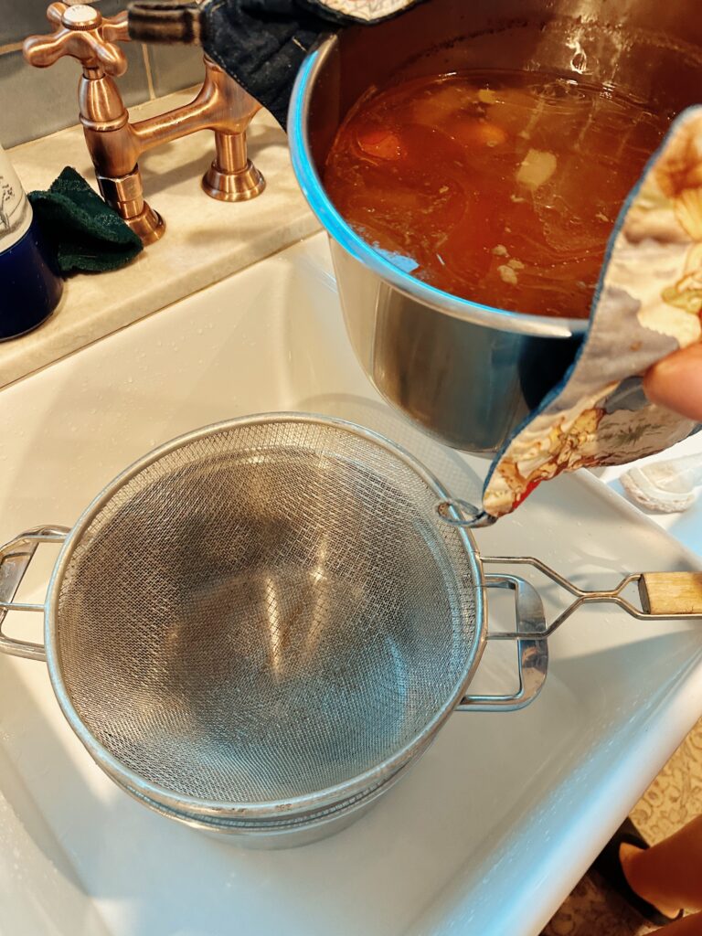 A colander over a metal pot in a sink with the instant pot of broth hovering over it.
