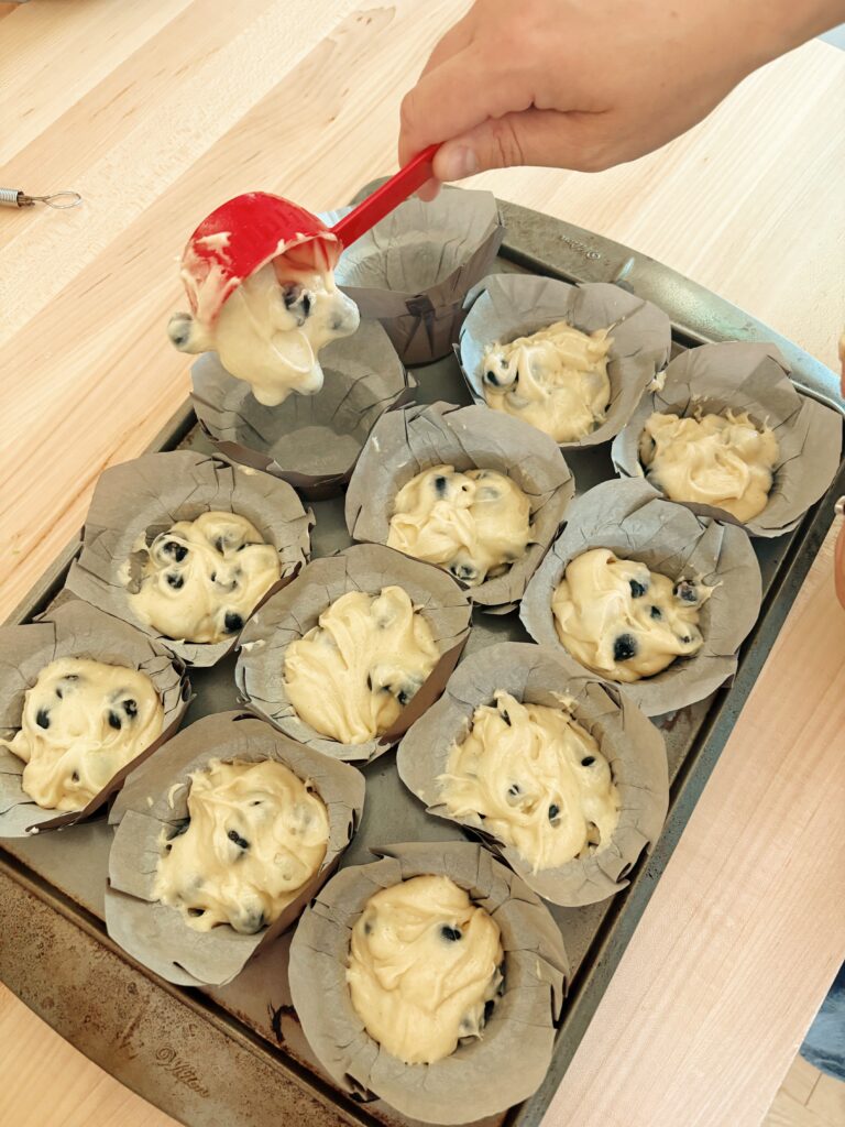 Muffin liners in a 12 muffin tin. The person is pouring a 1/4 cup of muffin better into each liner. 