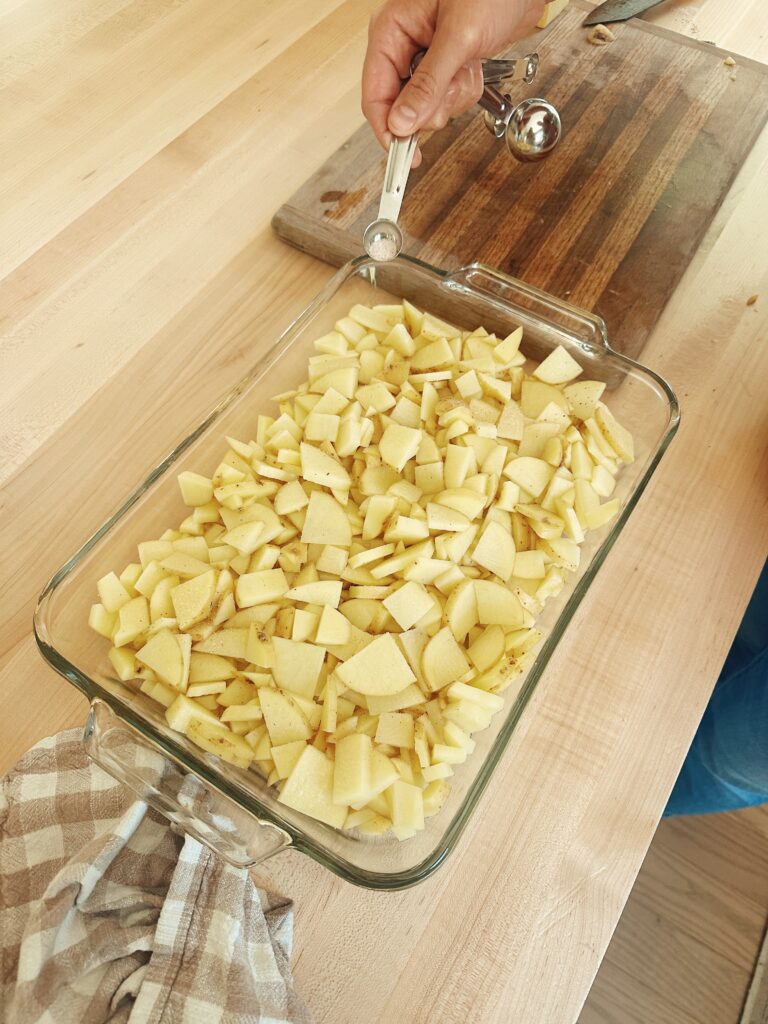 The sliced potatoes have been placed in a glass casserole dish. The person is sprinkling half of the alt and pepper over them.