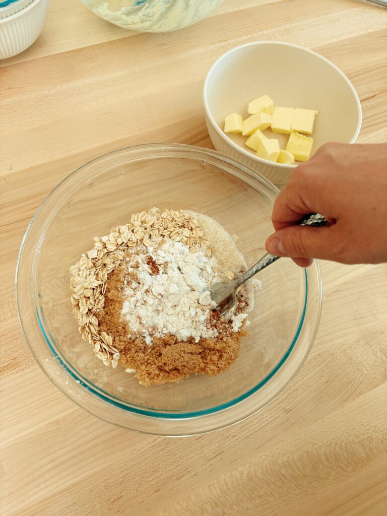 In a new glass bowl the person is combining the ingredients for the muffin crumble. Including: rolled oats, flour, brown sugar, and granulated sugar. 